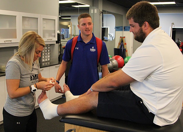 Foster gets his ankles taped by athletic trainer Lauren Askevold; Foster’s teammate Jackson Dick of Tallahassee, Fla., a dual-sport student-athlete in soccer and football, stops to chat.