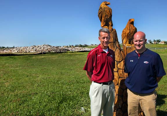 Joplin High School athletic director Jeff Starkweather (left) along with assistant athletic director Bruce Vonder Haar stand recently in front of what remains of the campus after it was destroyed by a tornado on May 22, 2011. The tree trunk behind them was left partially standing among a grove of trees that use to be in front of the school. An unknown person carved the artwork in honor of the school's mascot - the Eagles.  
