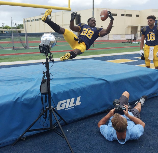 Photographer Stuart Browning (laying on the ground) takes a photo of St. Thomas Aquinas running back James Charles leaping over a track mat.